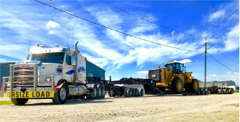 A large truck hauling a massive tractor down a dirt road.